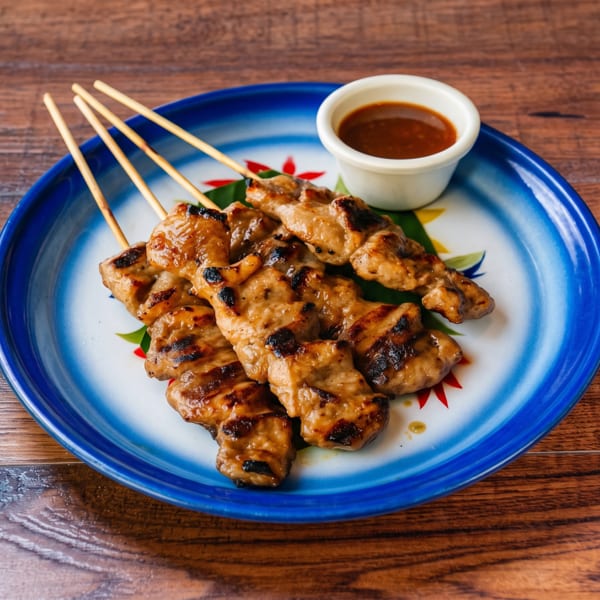 Pork satay skewers with a dipping sauce on a decorative blue and white plate.