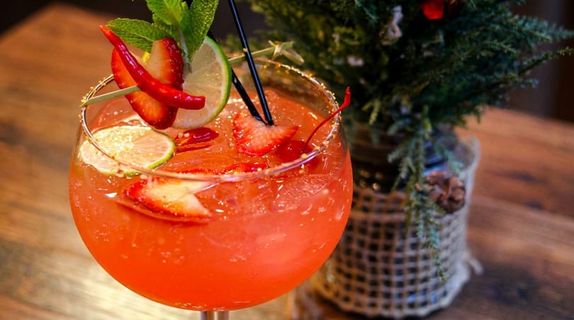 A vibrant red cocktail garnished with strawberries, lime, mint, and chili peppers, next to a small Christmas tree.