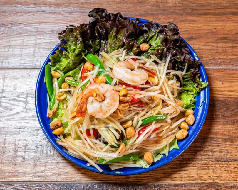Thai papaya salad with shrimp, peanuts, green beans, and lettuce on a blue plate.