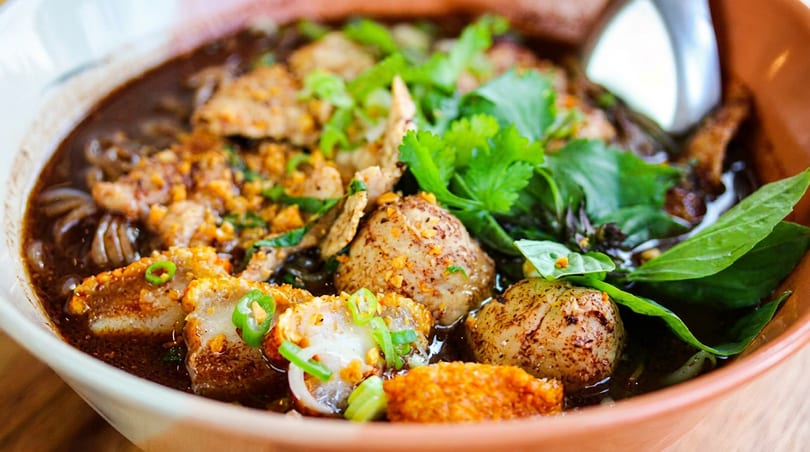 Close-up of a bowl of dark broth noodle soup with meat, herbs, and crispy toppings.