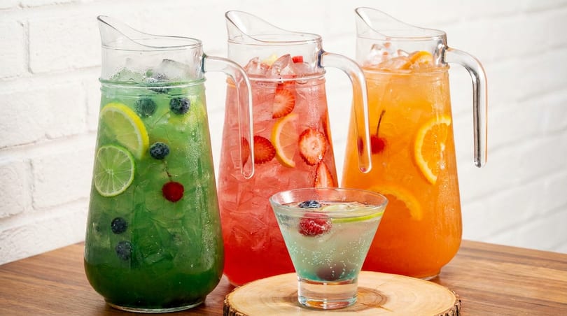 Three pitchers of colorful fruit-infused drinks and one glass on a wooden surface.