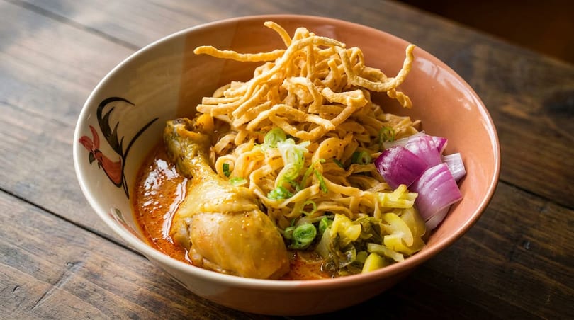 A bowl of Khao Soi with chicken, crispy noodles, red onions, and pickled greens on a wooden table.