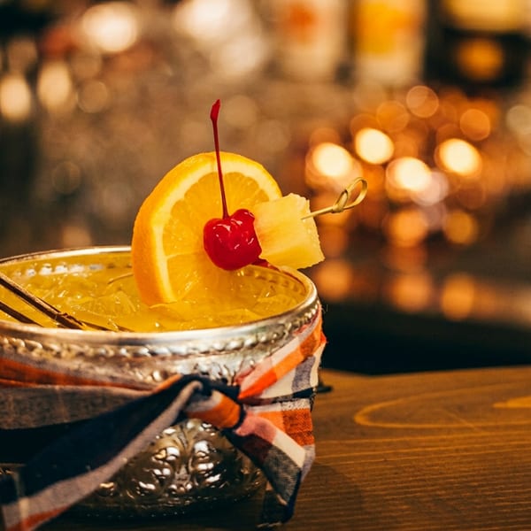 A vibrant yellow cocktail in an ornate silver cup, garnished with orange, cherry, and pineapple.