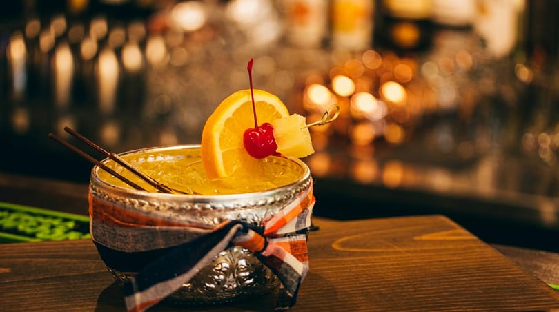 A vibrant yellow cocktail in an ornate silver cup, garnished with orange, cherry, and pineapple.