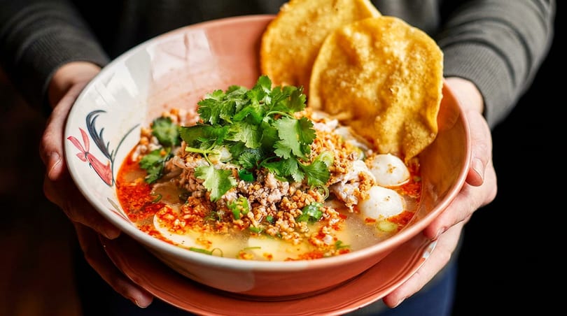 Hands holding a bowl of Thai noodle soup with minced pork, fish balls, and cilantro.