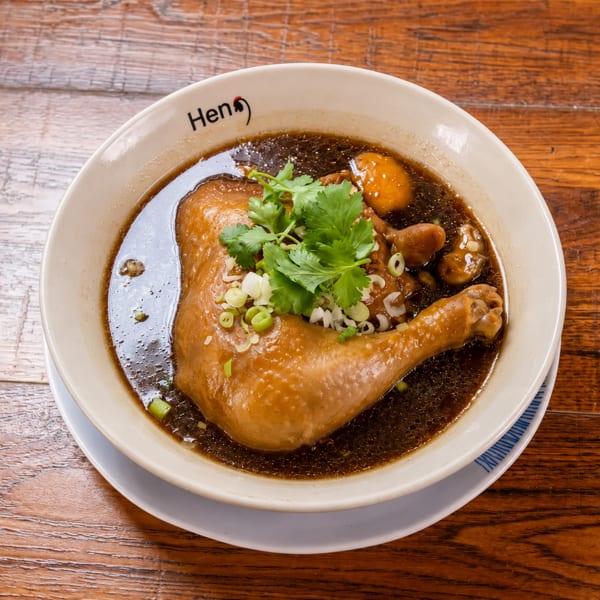Chicken leg in dark broth with cilantro and green onions in a "Hen" branded bowl on a wooden table.