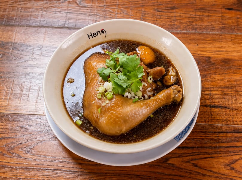 Chicken leg in dark broth with cilantro and green onions in a "Hen" branded bowl on a wooden table.