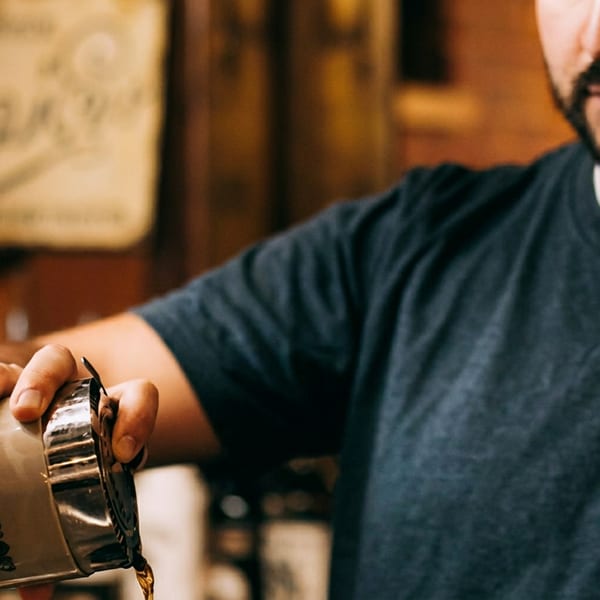 A bartender pours a drink from a silver Patron cocktail shaker, liquid streaming into a glass.