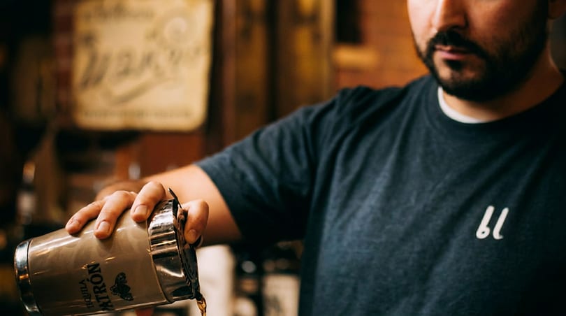 A bartender pours a drink from a silver Patron cocktail shaker, liquid streaming into a glass.