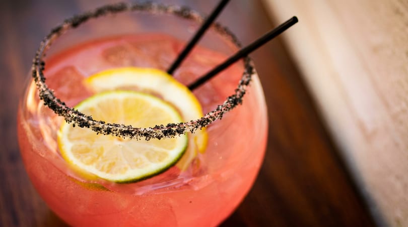 Pink cocktail with citrus slices, ice, two black straws, and a dark sugar-salted rim.