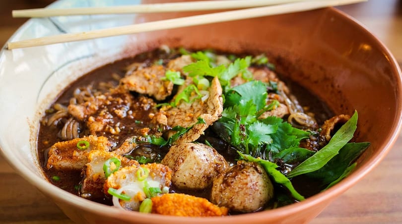 Close-up of a bowl of Thai boat noodles with pork, meatballs, and fresh herbs.