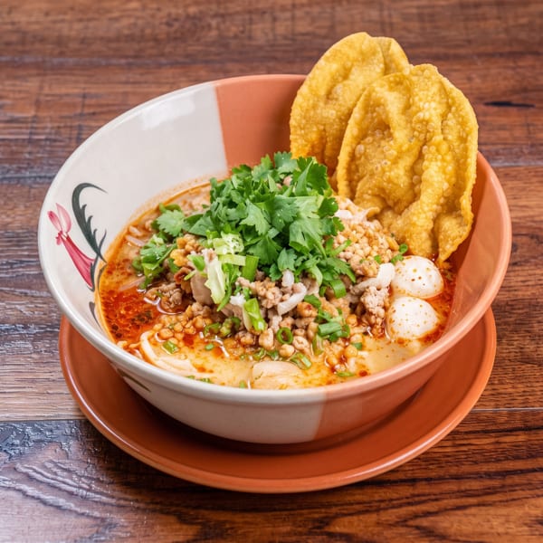 Decorative bowl of noodle soup with minced meat, fish balls, cilantro, and crispy wontons on wood.