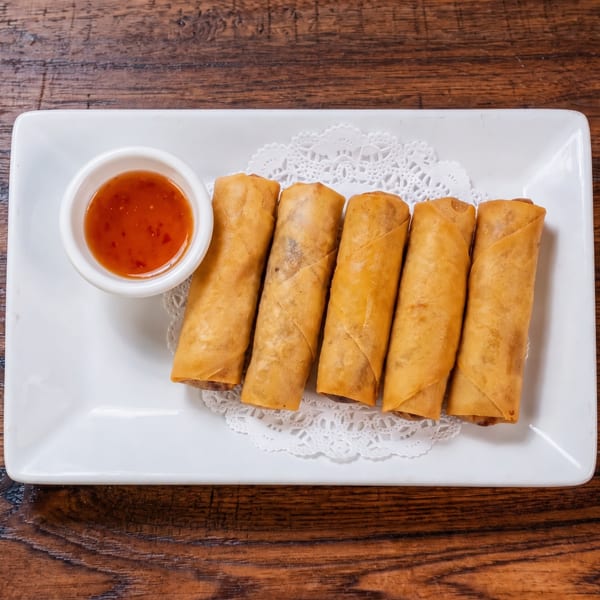 Six crispy spring rolls with sweet chili dipping sauce on a white rectangular plate.