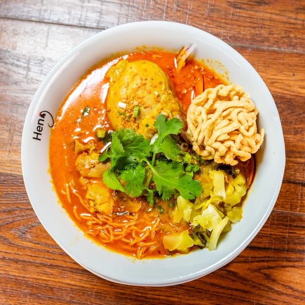 Heng's Khao Soi with chicken, crispy noodles, cilantro, and pickled vegetables on a wooden table.