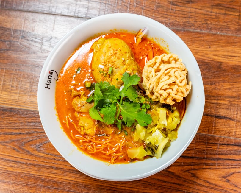 Heng's Khao Soi with chicken, crispy noodles, cilantro, and pickled vegetables on a wooden table.