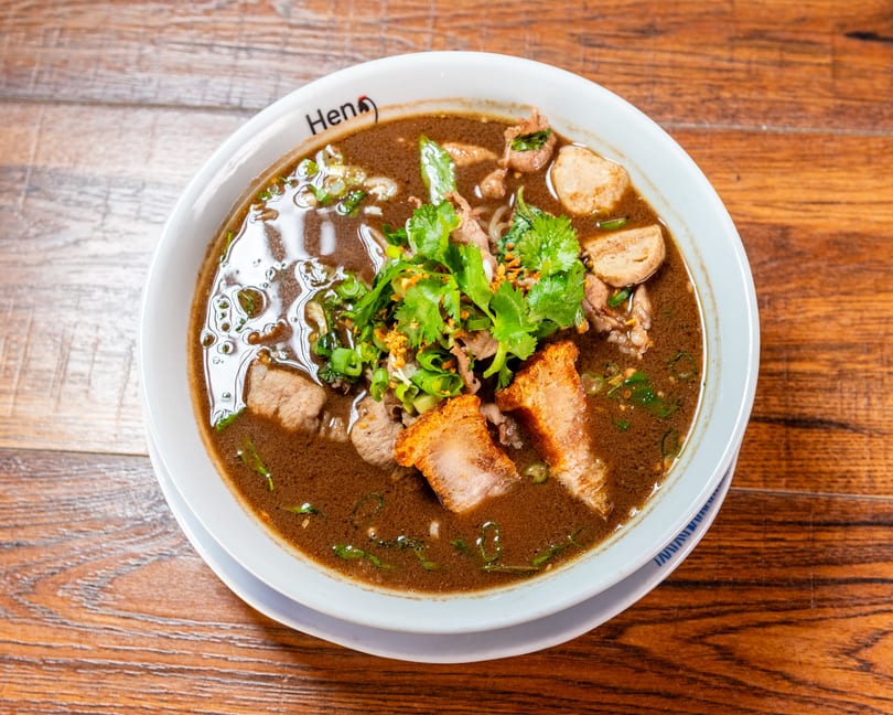 Dark broth noodle soup with meat, meatballs, crispy pork, cilantro, and green onions on a wooden table.