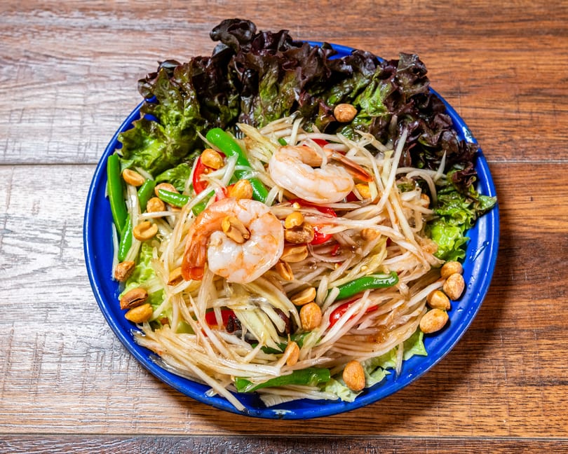 Fresh papaya salad with shrimp, peanuts, green beans, and lettuce on a blue plate.