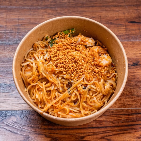Pad Thai noodles with shrimp and crushed peanuts in a takeout bowl on a wooden table.